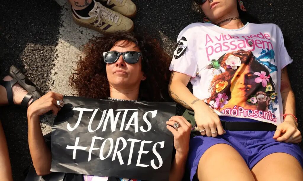 Duas mulheres deitadas no aslfato durante mobilização política. Uma segura um cartaz escrito "Juntas somos mais fortes". A outra exibe uma camiseta. Foto: Rovena Rosa / Agência Brasil